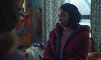 Movie still from “The Silent Twins” (2022), directed by Agnieszka Smoczynska – A woman in a pink jacket sitting in a room; Close Up shot, Over the shoulder angle