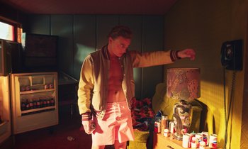 Movie still from “The Silent Twins” (2022), directed by Agnieszka Smoczynska – A man standing in front of a table with a bottle; Medium shot, Low angle