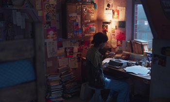 Movie still from “The Silent Twins” (2022), directed by Agnieszka Smoczynska – A person sitting at a desk in front of a bunch of papers; Wide shot, High angle