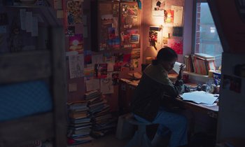 Movie still from “The Silent Twins” (2022), directed by Agnieszka Smoczynska – A person sitting at a desk in front of a bunch of papers; Wide shot, High angle