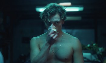 Movie still from “The Silent Twins” (2022), directed by Agnieszka Smoczynska – A shirtless man holding a piece of food in front of his face; Medium shot, Low angle