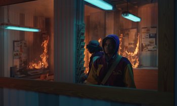 Movie still from “The Silent Twins” (2022), directed by Agnieszka Smoczynska – A man wearing a hooded jacket standing in front of a fire; Medium shot, Low angle