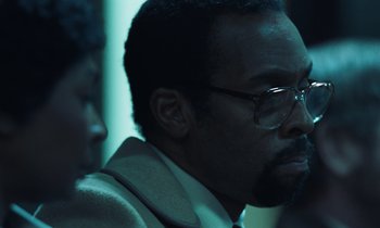 Movie still from “The Silent Twins” (2022), directed by Agnieszka Smoczynska – A person wearing a suit and glasses; Close Up shot, Over the shoulder angle