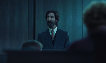 Movie still from “The Silent Twins” (2022), directed by Agnieszka Smoczynska – A man in a suit and tie in front of a group of people; Medium shot, Over the shoulder angle