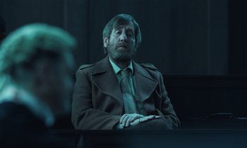 Movie still from “The Silent Twins” (2022), directed by Agnieszka Smoczynska – A man in a suit and tie sitting at a table; Medium shot, Over the shoulder angle