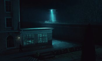 Movie still from “The Silent Twins” (2022), directed by Agnieszka Smoczynska – A building in the middle of the night with a light shining on it; Extreme Wide shot, High angle