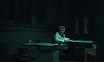 Movie still from “The Silent Twins” (2022), directed by Agnieszka Smoczynska – A man sitting at a table in a dark room; Wide shot, Low angle