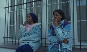 Movie still from “The Silent Twins” (2022), directed by Agnieszka Smoczynska – Two young women sitting next to each other smoking cigarettes; Medium shot, Low angle