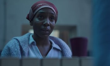 Movie still from “The Silent Twins” (2022), directed by Agnieszka Smoczynska – A woman wearing a head scarf looking at the camera; Close Up shot, Low angle
