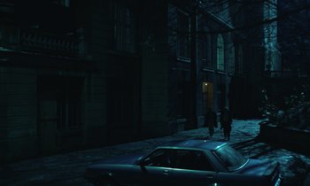 Movie still from “The Silent Twins” (2022), directed by Agnieszka Smoczynska – Two people walking down a street next to a parked car; Extreme Wide shot, High angle