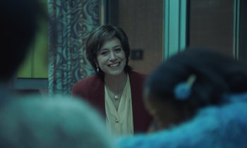 Movie still from “The Silent Twins” (2022), directed by Agnieszka Smoczynska – A woman smiles while talking to a group of people; Medium shot, Over the shoulder angle