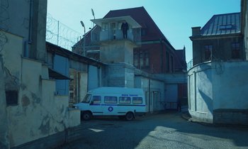Movie still from “The Silent Twins” (2022), directed by Agnieszka Smoczynska – A van parked in front of a building with a man standing on top of it; Extreme Wide shot, Low angle
