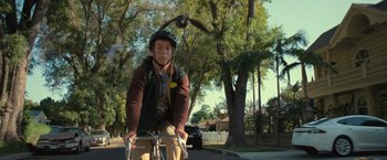 Movie still from “The Starling” (2021), directed by Theodore Melfi – A person riding a bike on a street near a tree; Medium shot, Over the shoulder angle