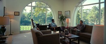Movie still from “The Starling” (2021), directed by Theodore Melfi – A man sitting on a couch in front of a large window; Wide shot, Over the shoulder angle