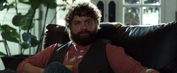 Movie still from “Due Date” (2010), directed by Todd Phillips – A bearded man sitting in a chair holding a drink; Medium shot, Over the shoulder angle