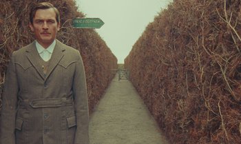 Movie still from “The Swan” (2023), directed by Wes Anderson – A man in a suit standing in front of bushes; Medium shot, Over the shoulder angle