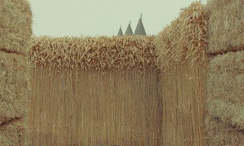 Movie still from “The Swan” (2023), directed by Wes Anderson – A wall made out of straw with three buildings in the background; Extreme Wide shot, Low angle