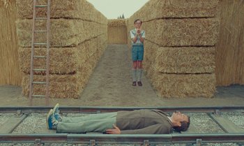 Movie still from “The Swan” (2023), directed by Wes Anderson – A man laying on hay bales next to another man standing on a train track; Wide shot, Overhead angle