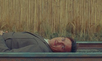 Movie still from “The Swan” (2023), directed by Wes Anderson – A man in a suit laying on the ground in front of a fence; Medium shot, Overhead angle