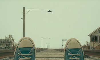 Movie still from “The Swan” (2023), directed by Wes Anderson – Two pairs of shoes sitting on the side of a road; Wide shot, Overhead angle