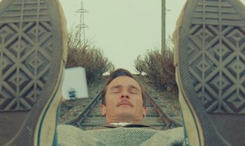 Movie still from “The Swan” (2023), directed by Wes Anderson – A man taking a picture of himself laying on a train track; Close Up shot, Overhead angle