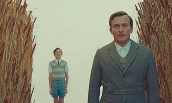 Movie still from “The Swan” (2023), directed by Wes Anderson – A man and a boy standing next to each other in front of tall grass; Medium shot, Low angle