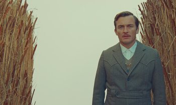 Movie still from “The Swan” (2023), directed by Wes Anderson – A man wearing a suit and tie standing in front of some tall grass; Medium shot, Low angle