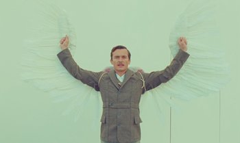 Movie still from “The Swan” (2023), directed by Wes Anderson – A man in a jacket and tie holding a pair of wings; Medium shot, Low angle