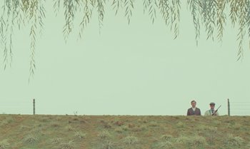 Movie still from “The Swan” (2023), directed by Wes Anderson – A man standing on top of a grass covered field; Extreme Wide shot, Low angle
