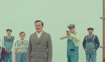 Movie still from “The Swan” (2023), directed by Wes Anderson – A group of men standing next to each other in front of a white wall; Medium shot, Low angle