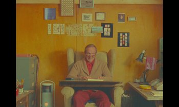 Movie still from “The Swan” (2023), directed by Wes Anderson – An older man sitting in a chair with a laptop; Wide shot, High angle