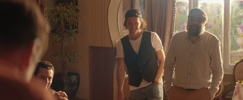 Movie still from “The Swimmers” (2022), directed by Sally El Hosaini – A young man in a black vest is smiling for the camera; Medium shot, Over the shoulder angle