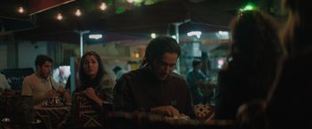 Movie still from “The Swimmers” (2022), directed by Sally El Hosaini – A man sitting in front of a table with other people in the background; Medium shot, Over the shoulder angle