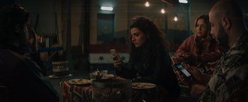Movie still from “The Swimmers” (2022), directed by Sally El Hosaini – A woman sitting at a table looking at her phone; Medium shot, High angle
