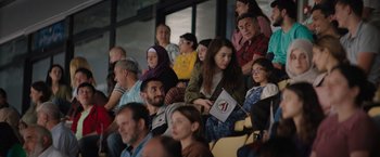 Movie still from “The Swimmers” (2022), directed by Sally El Hosaini – A group of people sitting in the stands at an event; Wide shot, High angle