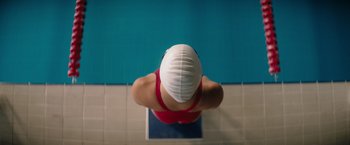 Movie still from “The Swimmers” (2022), directed by Sally El Hosaini – A woman in a red swimsuit and a white cap is in front of a swimming pool; Close Up shot, Overhead angle