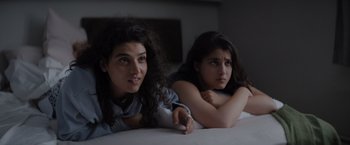 Movie still from “The Swimmers” (2022), directed by Sally El Hosaini – Two young women laying on a bed with a remote; Close Up shot, High angle