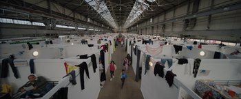 Movie still from “The Swimmers” (2022), directed by Sally El Hosaini – A group of people walking through a room with clothes hanging on them; Extreme Wide shot, Overhead angle