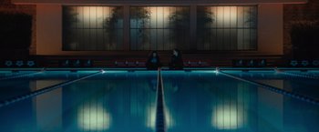Movie still from “The Swimmers” (2022), directed by Sally El Hosaini – Two people sitting in front of an indoor swimming pool at night; Extreme Wide shot, High angle