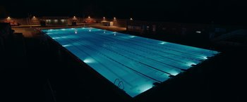 Movie still from “The Swimmers” (2022), directed by Sally El Hosaini – An empty swimming pool lit up at night time; Extreme Wide shot, High angle