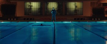 Movie still from “The Swimmers” (2022), directed by Sally El Hosaini – A man standing next to a swimming pool at night; Extreme Wide shot, High angle