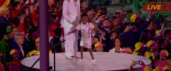 Movie still from “The Swimmers” (2022), directed by Sally El Hosaini – A young boy is walking on a stage while a crowd watches; Wide shot, Low angle