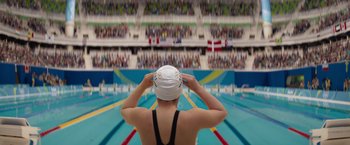 Movie still from “The Swimmers” (2022), directed by Sally El Hosaini – A woman in a swimming cap is standing in front of an audience; Extreme Wide shot, Overhead angle