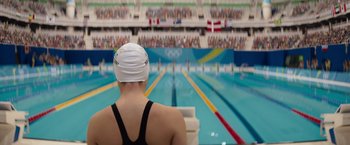 Movie still from “The Swimmers” (2022), directed by Sally El Hosaini – A woman in a swimming cap is sitting in front of an olympic swimming pool; Extreme Wide shot, High angle
