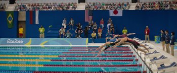 Movie still from “The Swimmers” (2022), directed by Sally El Hosaini – A group of people sitting on top of steps watching a swimming event; Extreme Wide shot, High angle