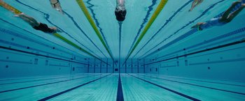 Movie still from “The Swimmers” (2022), directed by Sally El Hosaini – A person swimming in a pool under a blue sky; Extreme Wide shot, Overhead angle