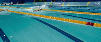 Movie still from “The Swimmers” (2022), directed by Sally El Hosaini – A woman swimming in a pool with a yellow and blue swimming lane; Extreme Wide shot, High angle