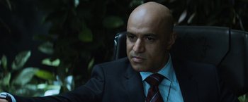 Movie still from “Elysium” (2013), directed by Neill Blomkamp – A man in a suit and tie sitting in a chair; Close Up shot, Over the shoulder angle