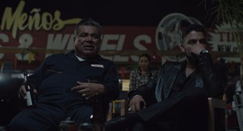 Movie still from “The Tax Collector” (2020), directed by David Ayer – A man sitting next to another man holding a drink; Medium shot, Over the shoulder angle