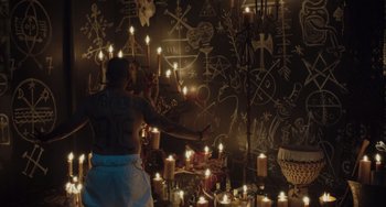 Movie still from “The Tax Collector” (2020), directed by David Ayer – A man standing in front of a wall covered in candles; Wide shot, High angle